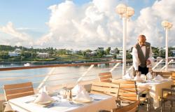 Azamara Quest - Azamara Club Cruises - čísník připravujíc jídelní stoly na venkovní terasa a v pozadí panorama luxusních vil a domků na zeleném pobřeží